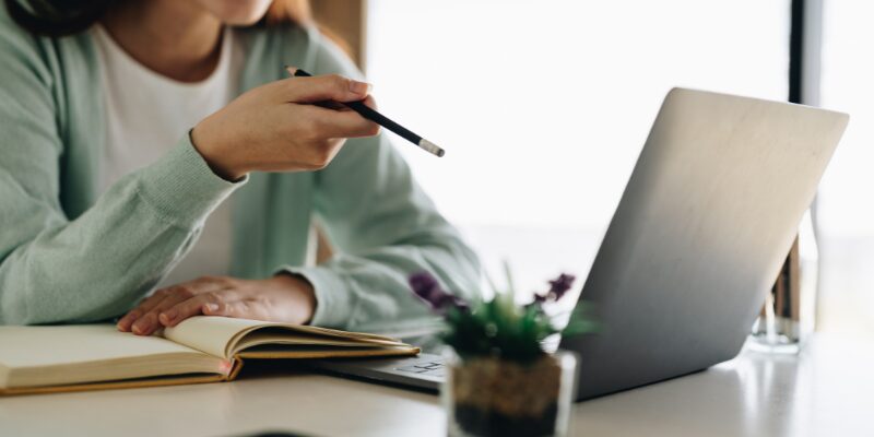 Donna sul tavolo con il computer davanti e con in mano una matita. Prende appunti sul quaderno.