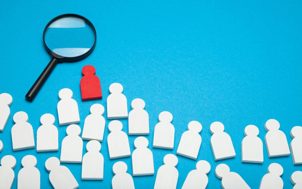 Gruppo di sagome stilizzate bianche su sfondo azzurro, tra cui spicca una figura rossa. Una lente di ingrandimento posta sopra di essa ne evidenzia la diversità rispetto alle altre, evocando il concetto di identificazione, diagnosi o individuazione di una caratteristica specifica in mezzo a un gruppo.