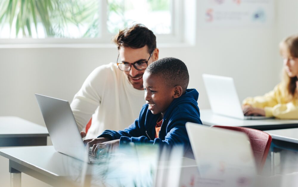 Insegnante con occhiali che sorride affiancando un bambino davanti a un laptop in un'aula scolastica luminosa. Sullo sfondo, un'altra bambina lavora al proprio computer. L'immagine evoca un momento di supporto individualizzato e apprendimento digitale.