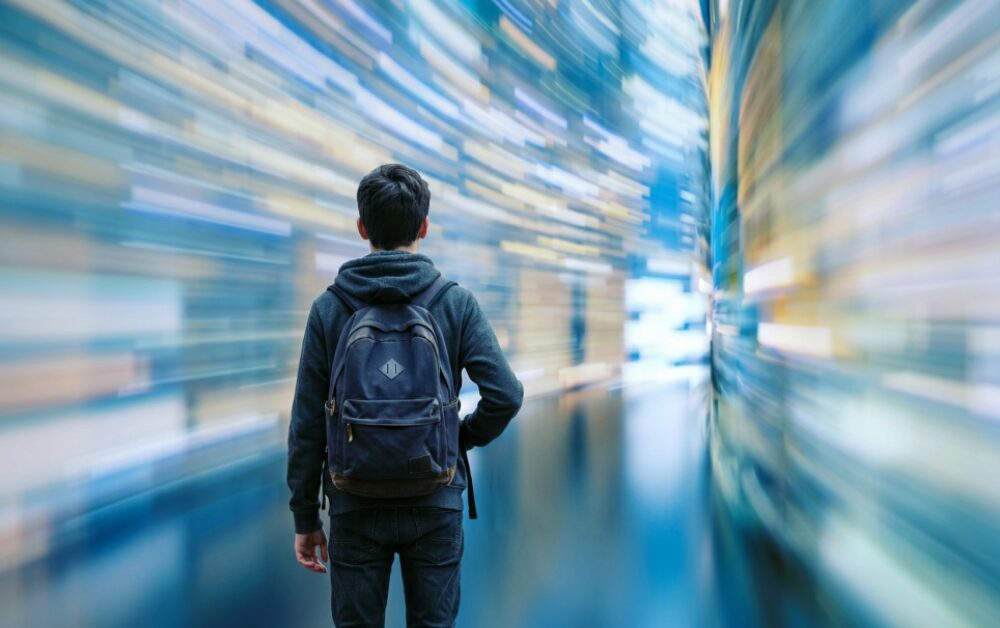 Ragazzo visto di spalle con uno zaino, in piedi al centro di un corridoio avvolto da luci colorate sfocate in blu, giallo e bianco che creano un effetto di velocità o immersione digitale, evocando il rapporto tra giovani e flusso di informazioni.