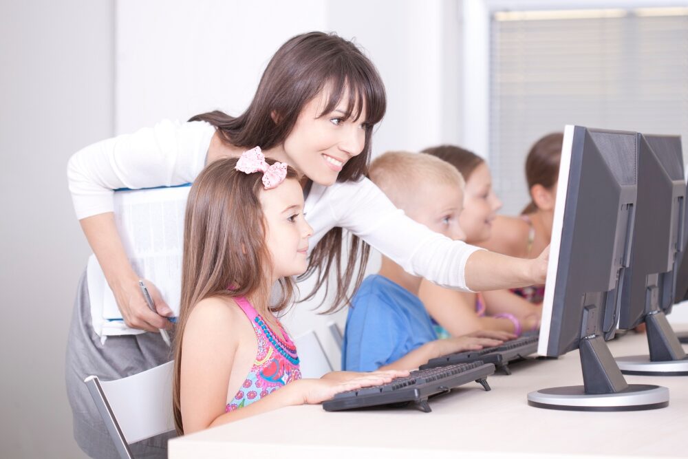 Insegnante sorridente che si china verso una bambina seduta davanti a un computer desktop in un'aula di informatica, indicando lo schermo per guidarla. Sullo sfondo, altri bambini lavorano ai propri computer in fila.