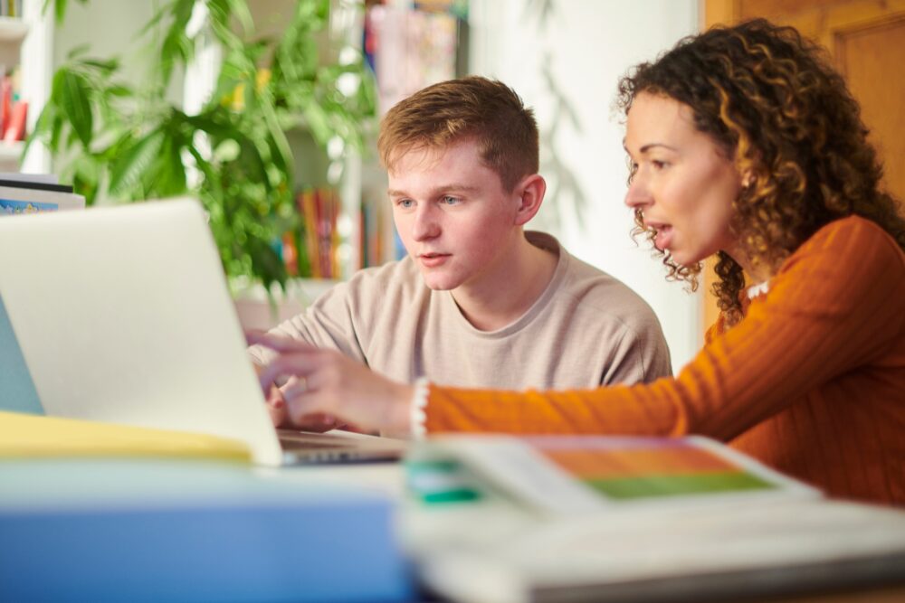Ragazzo adolescente seduto davanti a un laptop affiancato ad un tutor.