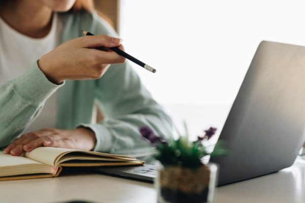 Donna sul tavolo con il computer davanti e con in mano una matita. Prende appunti sul quaderno.