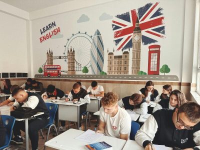 Aula scolastica con studenti adolescenti seduti ai banchi, intenti a studiare o svolgere esercizi. Sulla parete di sfondo un murale colorato con i simboli di Londra — il Tower Bridge, il London Eye, il Big Ben, un autobus rosso e una cabina telefonica — accompagnato dalla scritta 'Learn English'.