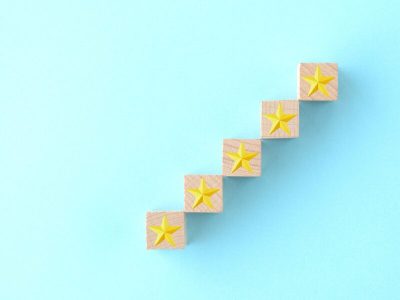 Cinque cubi di legno disposti a scalare dal basso a sinistra verso l'alto a destra, ciascuno con una stella dorata impressa sulla superficie, su sfondo azzurro. L'immagine evoca una progressione crescente e il concetto di valutazione o miglioramento graduale.