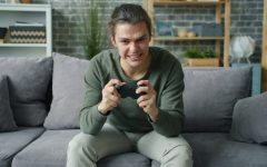 Ragazzo adolescente seduto sul divano di casa con un'espressione di intensa concentrazione, che stringe un controller da videogioco con entrambe le mani. Sullo sfondo, una stanza con scaffali e piante.