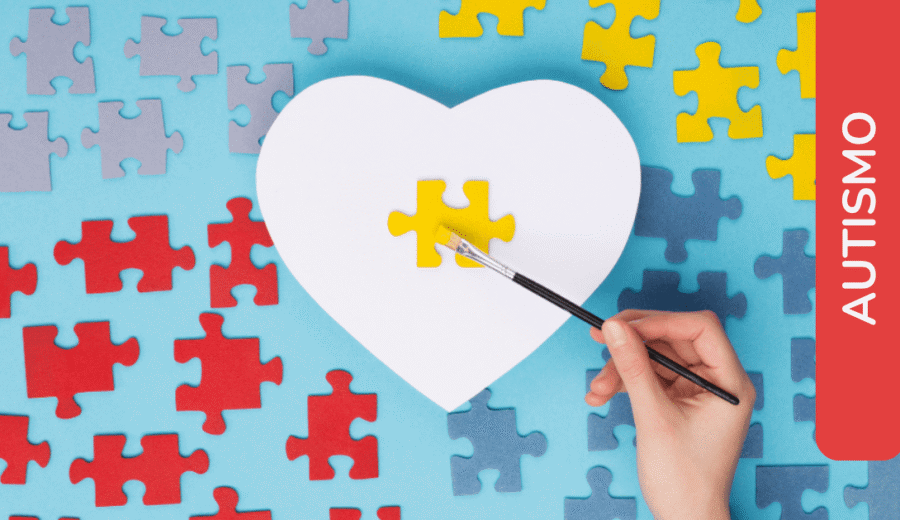 Mano che dipinge un pezzo di puzzle giallo su un cuore bianco, circondato da pezzi colorati. Etichetta: "Autismo".