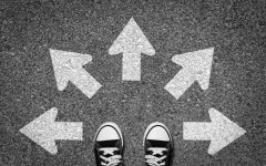 Vista dall'alto di un paio di sneakers nere su asfalto, circondate da cinque freccie bianche dipinte che puntano in direzioni diverse — sinistra, destra, alto, e diagonali — suggerendo molteplici possibilità o percorsi tra cui scegliere.