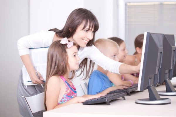 Insegnante sorridente che si china verso una bambina seduta davanti a un computer desktop in un'aula di informatica, indicando lo schermo per guidarla. Sullo sfondo, altri bambini lavorano ai propri computer in fila.