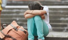 Foto di una ragazzina con la testa fra le mani in atteggiamento di tristezza, seduta sui gradini di una scalinata, con lo zaino abbandonato a fianco