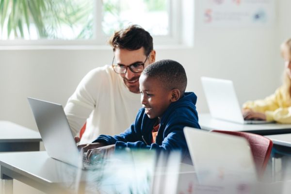 Insegnante con occhiali che sorride affiancando un bambino davanti a un laptop in un'aula scolastica luminosa. Sullo sfondo, un'altra bambina lavora al proprio computer. L'immagine evoca un momento di supporto individualizzato e apprendimento digitale.