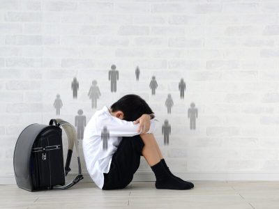 Bambino in età scolare seduto a terra con la testa tra le braccia, in atteggiamento di isolamento o tristezza. Accanto a lui uno zaino scolastico. Sullo sfondo, sagome stilizzate di persone che lo circondano, evocando ansia sociale o esclusione.