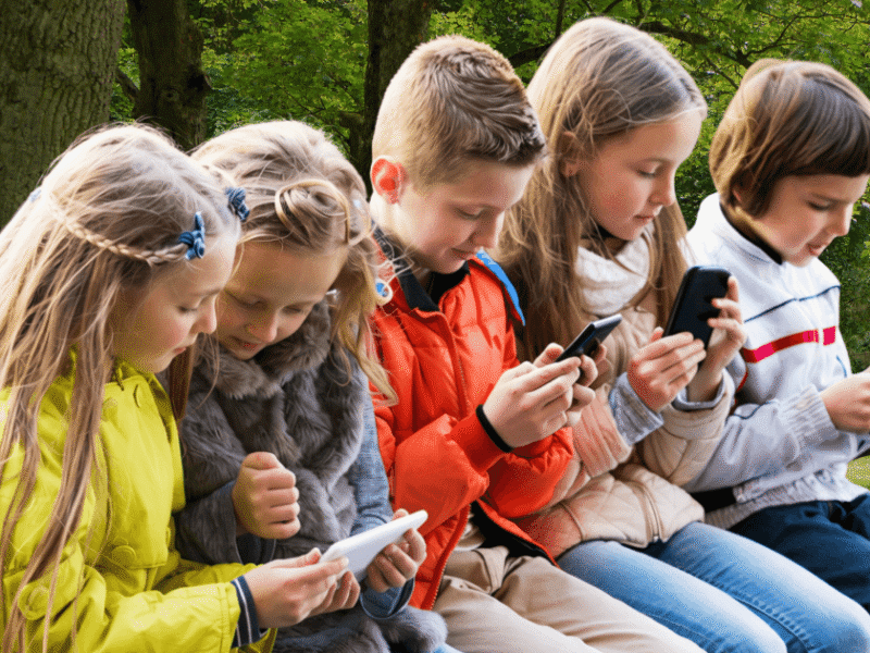 Cinque bambini seduti su una panchina in un parco, ognuno con in mano uno smartphone o tablet.