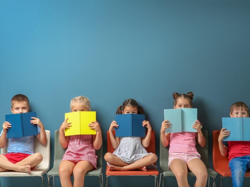 Cinque bambini in età prescolare seduti in fila su sedie colorate davanti a una parete azzurra, ciascuno con un libro aperto che copre parzialmente il volto. I libri sono di colori diversi — blu, giallo, azzurro — in un'atmosfera giocosa legata alla lettura.