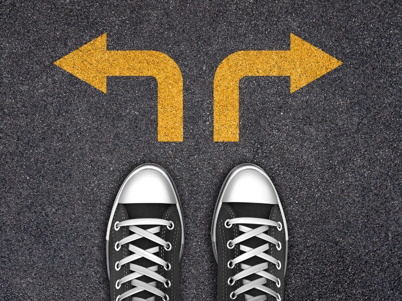 Vista dall'alto di un paio di sneakers nere su asfalto, davanti a due frecce gialle dipinte che indicano direzioni opposte, sinistra e destra, simbolo di una scelta o di un bivio.