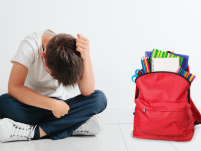 Bambino seduto a terra con la testa tra le mani, accanto a uno zaino rosso pieno di materiale scolastico.