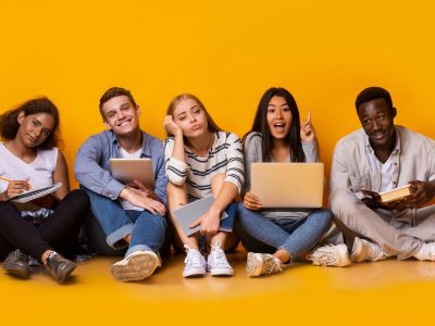 Cinque ragazzi adolescenti di diverse etnie seduti a terra davanti a uno sfondo giallo, con espressioni variegate — curiosità, noia, entusiasmo, sorriso. Ognuno tiene in mano uno strumento di studio diverso: un quaderno, un tablet, un laptop e un libro.