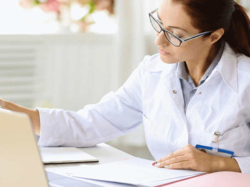 Professionista sanitaria in camice bianco davanti a un laptop, con penna in mano e documenti sul tavolo.
