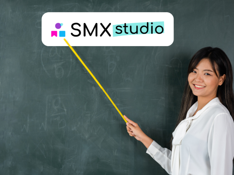Insegnante sorridente davanti a una lavagna che indica con un puntatore il logo di SMX Studio.