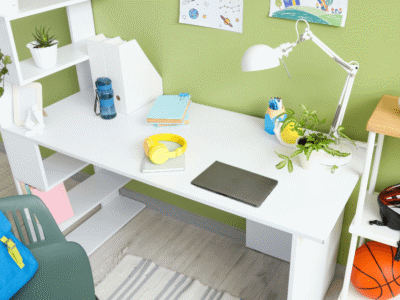 Scrivania bianca per bambini in una stanza verde, con laptop, cuffie gialle, zaino blu, piante e pallone da basket.